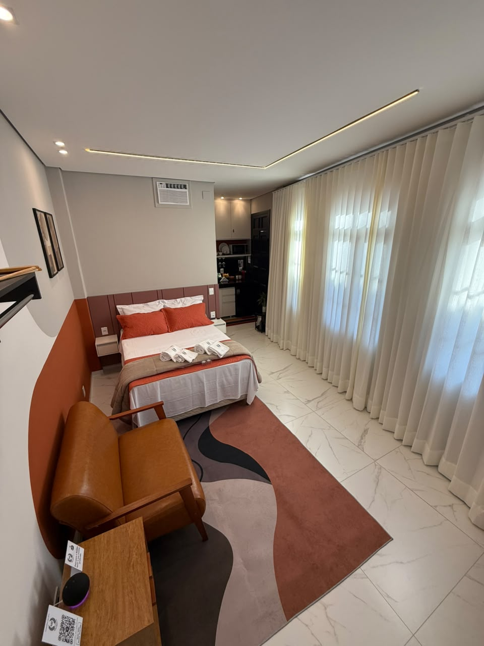 Studio amplo da Suíte Horto IV com cama de casal, poltrona de couro, tapete e cortinas até o teto