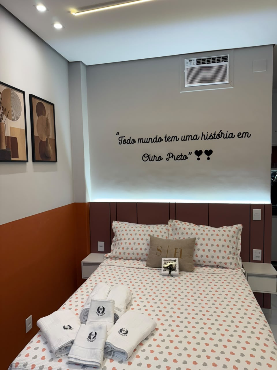 Quarto com cama de casal, roupa de cama de corações e a frase Todo mundo tem uma história em Ouro Preto na parede